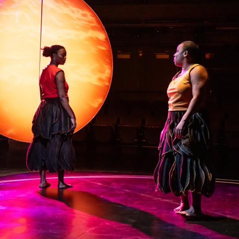 In front of a sun-like orb on a stage, two Black women dance in warm tank tops and billowing grey skirts.