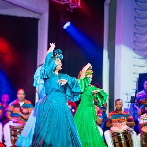On a stage, four women in monochromatic but bright colored costumes perform. They wear headwraps that match their dresses.