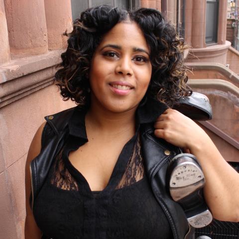 A brown woman holds shoes over her shoulder and has curly dark hair.