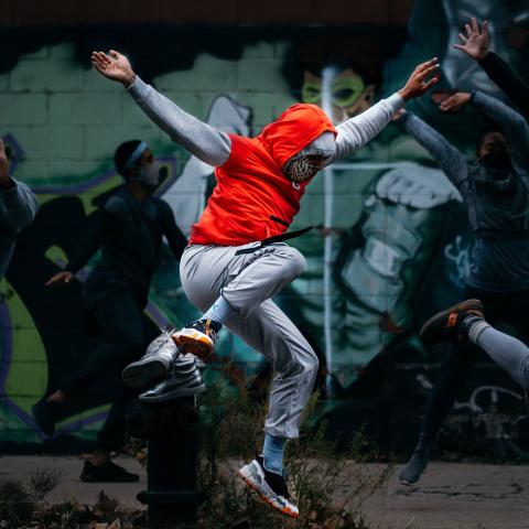 On a street and in front of a mural, a dancer throws their arms and leg up while wearing an orange hoodie.