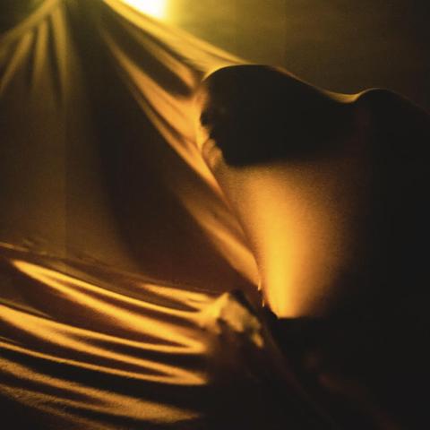 Under a spotlight, a person spreads golden satin over their face and stretches it with their hands.