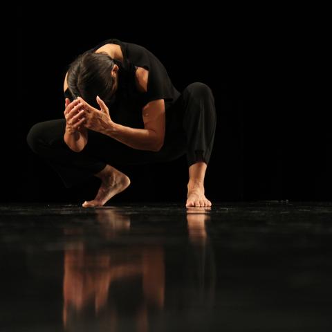 In a black background with body angled frontward and slightly towards the left, a Native woman dancer with medium brown skin, dark brown straight hair crouches in a semi-squat position with a curved back, elbows bent, and criss-crossed hands slightly covering the top of her head facing downward. One foot is flat on the ground while the other’s heel is raised. She wears black pants and a sleeveless top exposing both arms and shoulders. The black shiny flooring reveals a blurred reflection of her body.