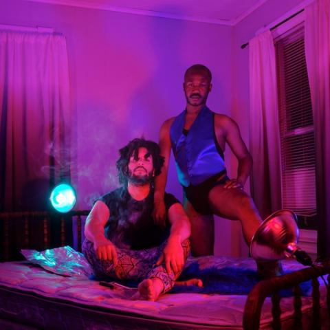 Two performers in a set with dim pink lighting. One sits on a bed; the stands behind with one leg extended on to the bed.
