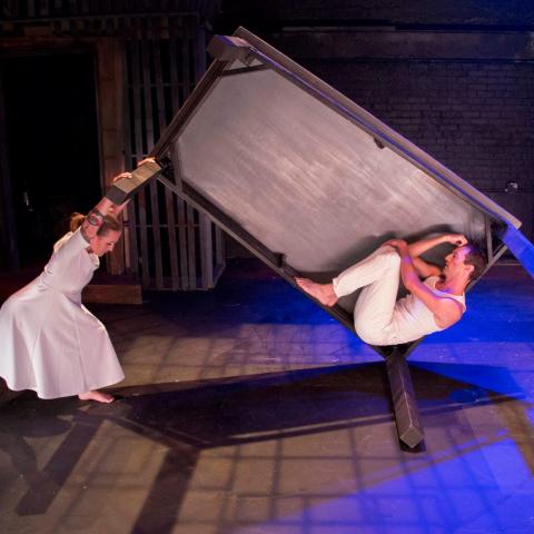 A white woman pushes a table on it's side that a white man sits inside the underside of.