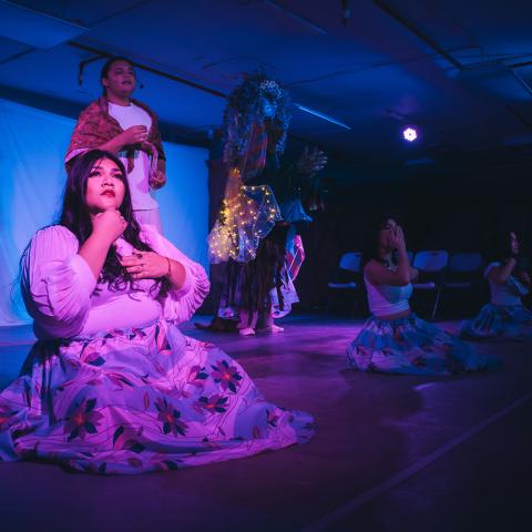 On a stage, a women kneels and holds herself in purple light. Behind her, more performers in the blue shadows.