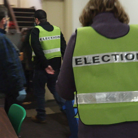 Folks, in vests that read "elections" on the back, walk towards a stairwell.
