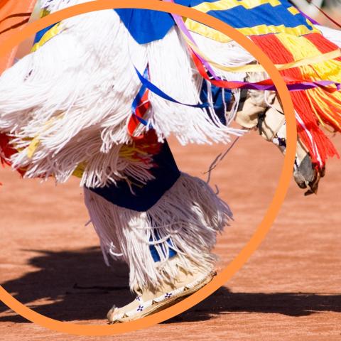 An orange outline of a sphere overlaid on an Indigenous pow-wow dancer’s shoes and dress.