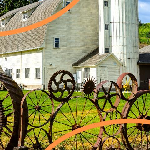 two orange curved lines overlay a large white barn with a sculpture made of rusty wheels in the foreground. 