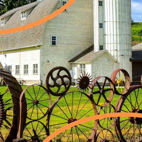 two orange curved lines overlay a large white barn with a sculpture made of rusty wheels in the foreground. 
