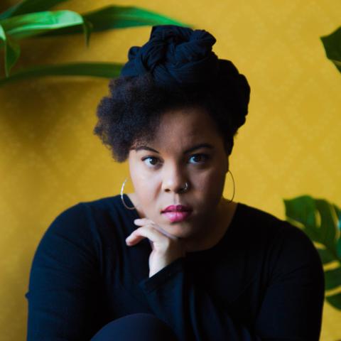 A Black woman poses in front of a yellow wall and bright green plants. She leans on her hand and wears gold hoops and all black.