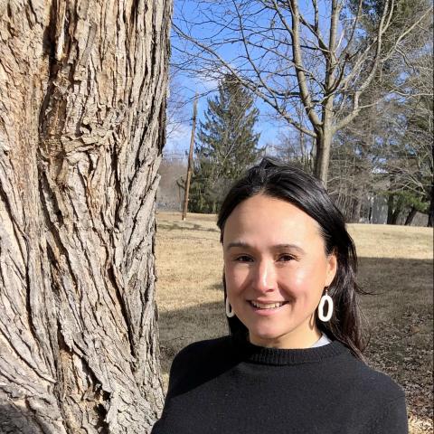 Brandi smiles next to a tree. She has light skin and black hair. She wears round white earrings.