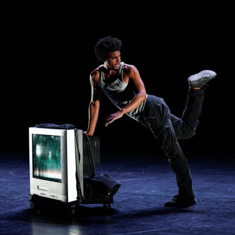 A performer kicks up a foot while leaning against a television box on a dark stage.