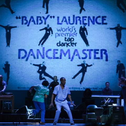 On a stage, with a projection that reads "Baby" Laurence works pefromed by a tap dancer dancemaster, three dancers tap in front of a live band.