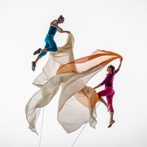 In a white void, two dancers (in brightly colored costumes) dance with orange and beige fabric.