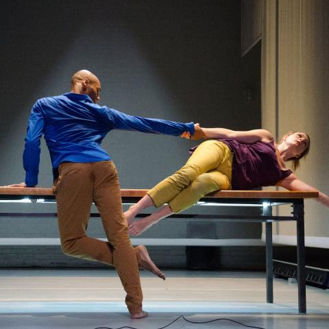 A Black man and a white woman perform over a table and under a spotlight,