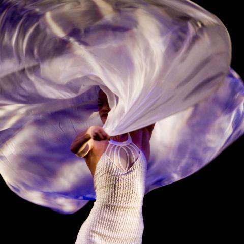 a woman in a white dress swirling purple fabric above her head and shoulders, partially obscuring her face. The backdrop is black.