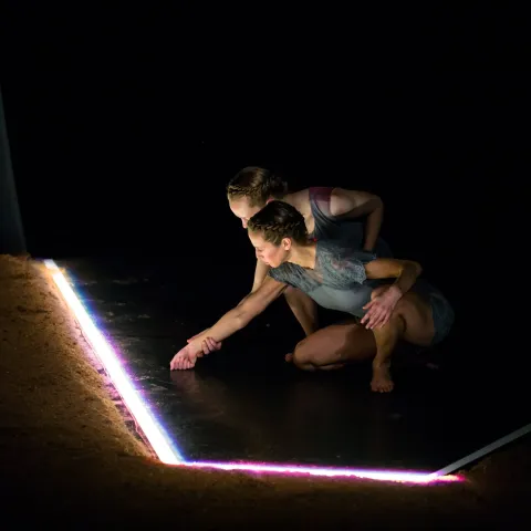 Two performers crouch by a bright tube of light and inspect it.