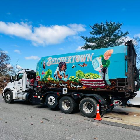 On the side of a large container on the back of a dump truck, a mural of kids watering plants. Monarch butterflies flutter. It reads behind them, "Belchertown," over a shade of blue found in the sky above.