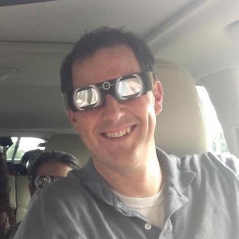 Man in his car with a child in the seat behind him, smiles in sunglasses.