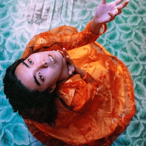 From above, a person dressed in orange is shown looking upwards while standing on a floral patterned blue floor.