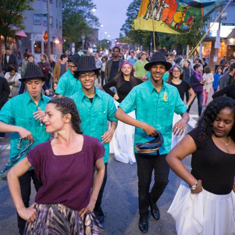 People dance on the street in Boston's Latin Quarter. Men in matching teal tees and black hats dance behind the first row of people.