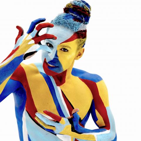 In a white space and covered in body paint, a person with a top bun touches their forehead.