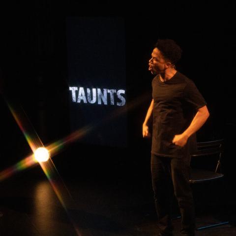 On a stage, a man of color stands before the word "Taunts." He is in all black and the stage is dimly lit.