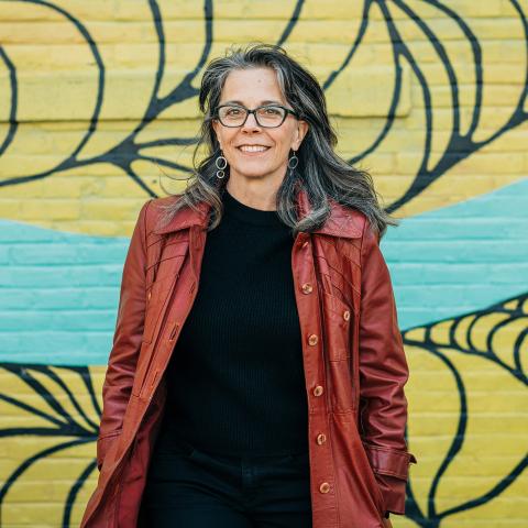 Britt is a white woman in a brown leather jacket. She poses in front of a mural.