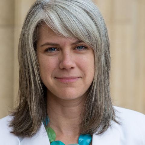 A woman with medium length hair wearing  a white blazer and a chunky blue and green necklace.