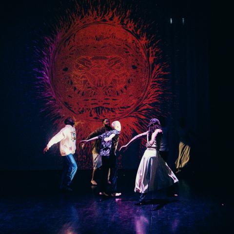 Four dancers perform in front of a red orb projected behind them.