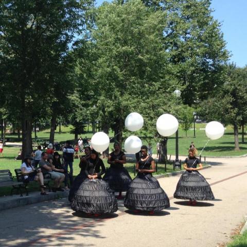 Five dancers in black dresses with white balloons slowly walk through the commons.