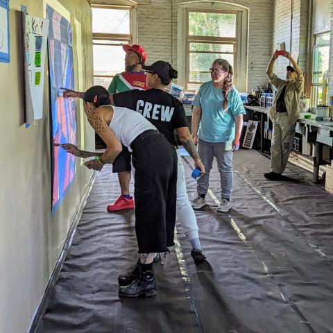 Four folks paint on a wall while two folks look on, one takes a photo on their phone.