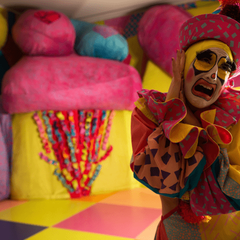 A drag queen dressed like a clown stands in front of a colorful background.