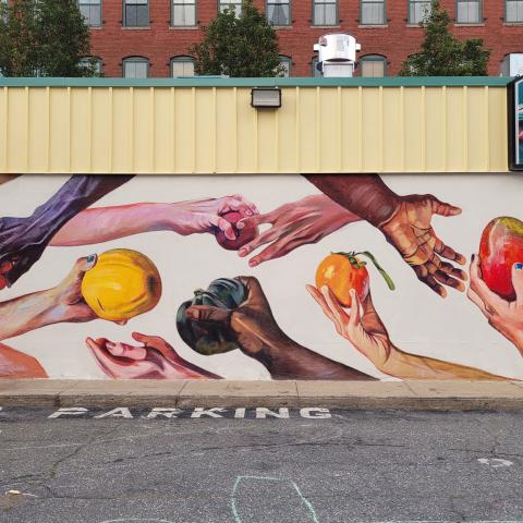 A mural on an external wall of a grocery store; on a white background,  the mural depicts hands of various skin tones handing pieces of fruit vegetables to others. "No Parking" is painted on the pavement in front of the mural.
