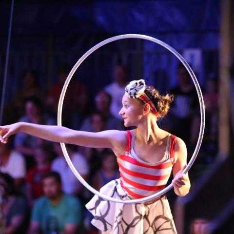 A woman holding a hula hoop with one hand and pointing her other arm through the hoop. She wears a red and white stripe tank top and a flouncy skirt, and goggles on her head.