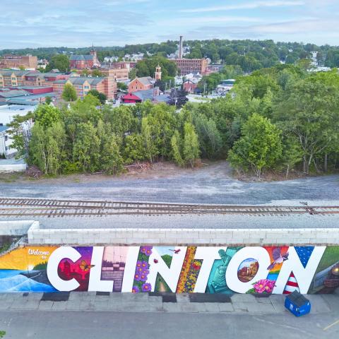 The mural, which spells out Clinton, sits on the side of a wall that creates a bridge for a train. The town of Clinton sits beyond the tracks with tall green trees and a bright blue sky.