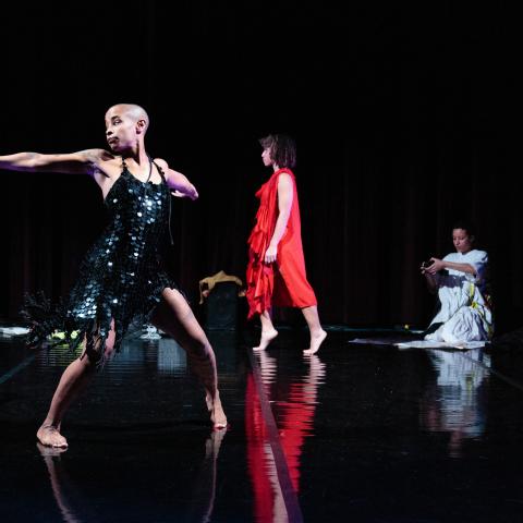 On a stage, a person in a Black flapper dress dances while a person in a red romper hops down and another person in a white dress kneels.