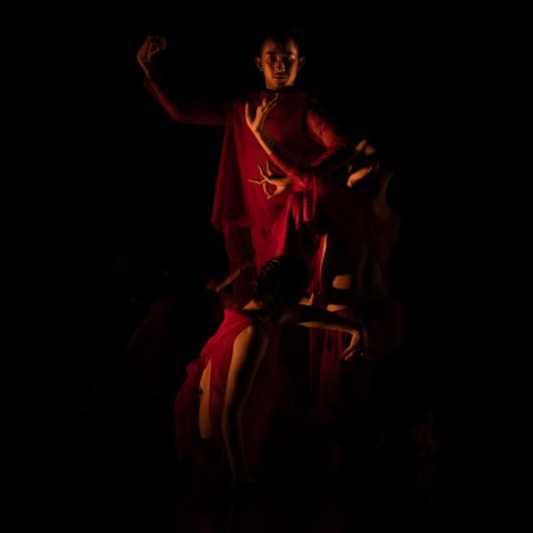 A dimly lit person, holds their hands up to their right and wears a long red cape.