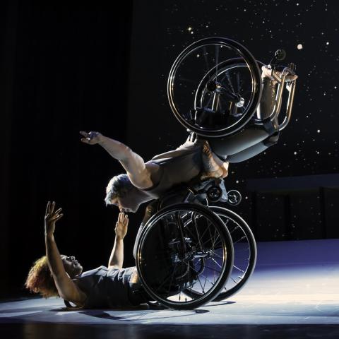 Two wheelchaired dancers in a starry setting. One is on their back on the ground and the other is somehow leaning on top of them without either leaving their chairs.