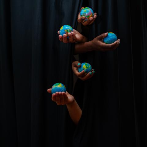 Five hands poke out from a set of black curtains, each one holds a miniature globe.