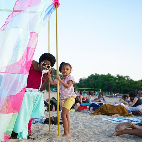 On the beach and within a wall of fabric, a woman holds a phone to her ear. A young girl stands next to her and holds onto the frame of the structure that holds up the wall of colorful fabric.