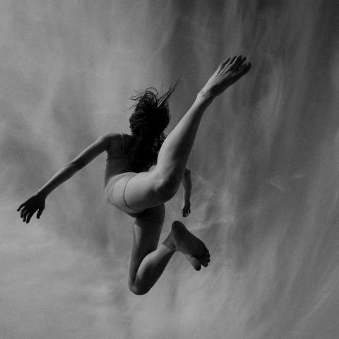 Black and white image, from underneath a person either leaps or is swimming. Stark contrast with their legs bent back and their hands reaching forward.