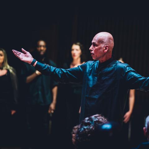 A bald man holds his arms out, in front of a group wearing all black.