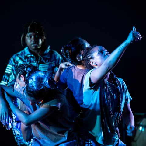 On a dark stage, with spotlights, four dancers of color look and point to the sky. They wear or appear in the light to wear blues and purples.