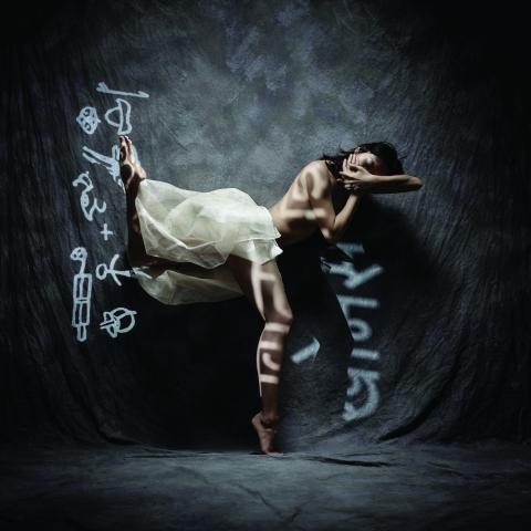 A topless woman dances with her arms over her chest in a curtained room with symbols projected over her.