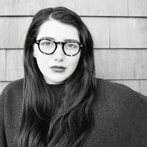 A woman with thick-framed glasses and long, brown hair poses in front of a building with wood siding.