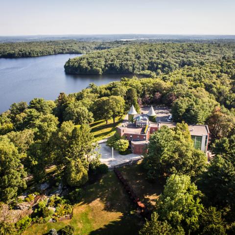 From overhead, by a pond in the woods, a brick building with dirt paths.