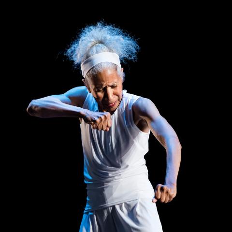 On a stage with blue and white spotlights, Diane wears white and holds her fists down and out in front of herself.