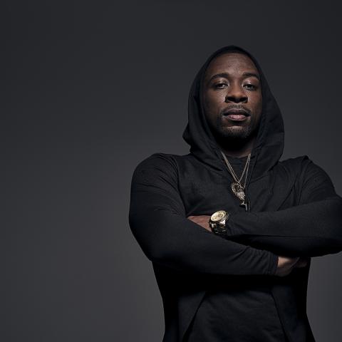 A black man crosses his arms. He poses in front of a gray backdrop and wears a black hoodie.