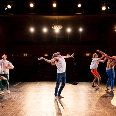 Five male dancers on a stage; one in the center with arms out and gazing upward; two pairs on either side making contact with each other. Costumes are casual street clothing